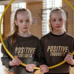 Twin girls in gym holding a hula hoop, wearing 'Positive Thoughts' sweatshirts.